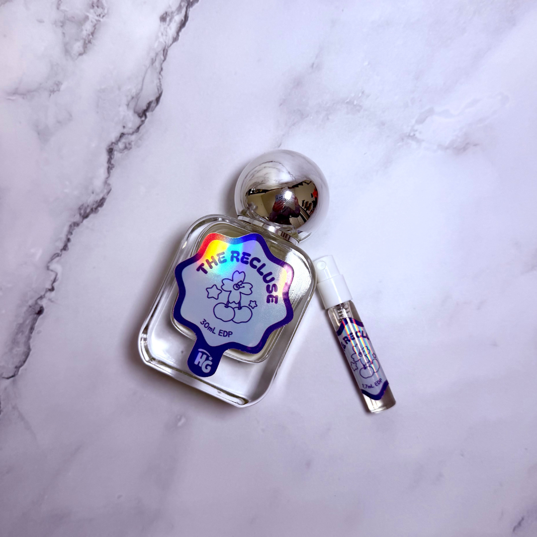 A clear glass perfume bottle with a reflective silver cap and a small matching spray vial, placed on a white marble surface. The bottle label reads 'THE RECLUSE' with a blue border and a rainbow holographic accent.