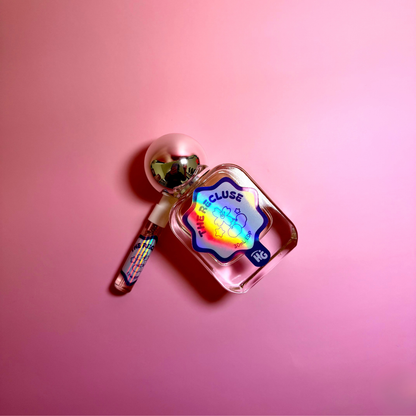 A small perfume bottle with a holographic blue label reading 'The Recluse' and a matching spray vial, photographed on a pink background.