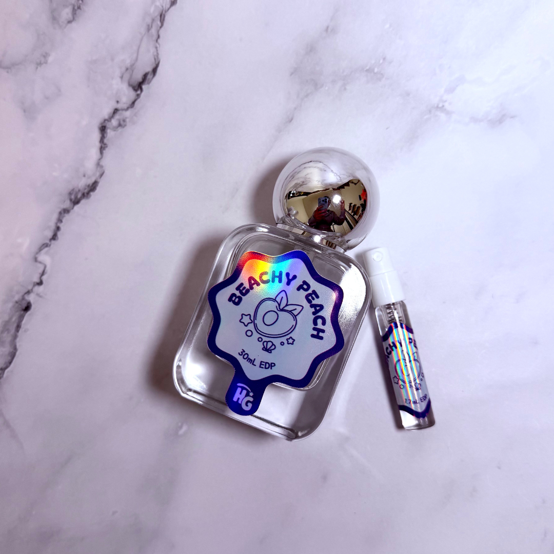 A clear glass perfume bottle labeled 'BEACHY PEACH' with a purple and holographic sticker, shown next to a small matching spray vial on a white marble surface.