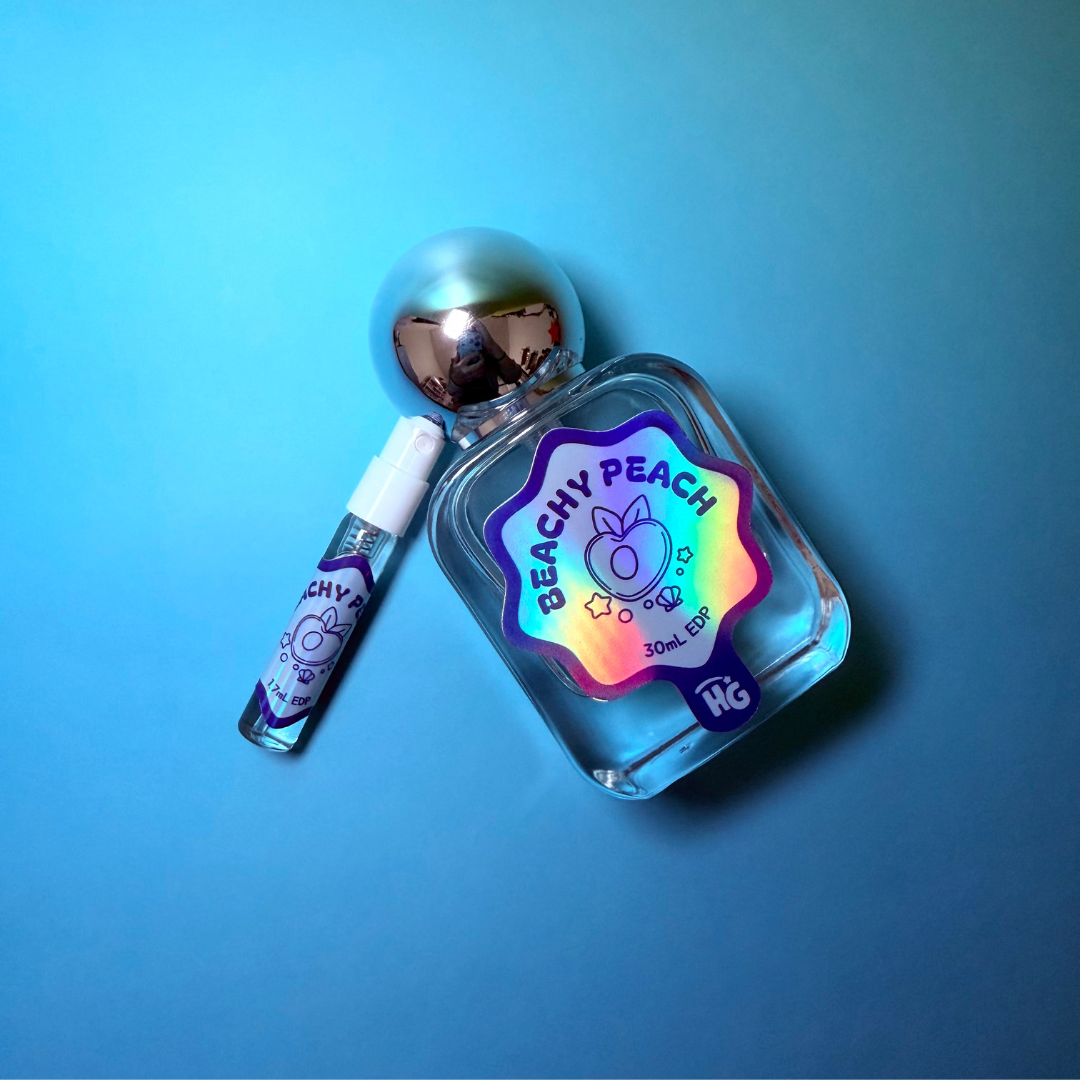 A small clear perfume bottle with a reflective metallic cap and a matching sample vial, both labeled 'BEACHY PEACH' on a blue gradient background.