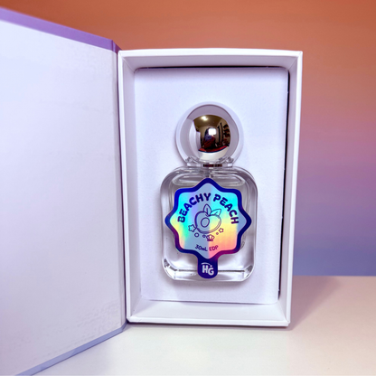 A clear glass perfume bottle with a metallic silver cap, presented inside an open white gift box. The bottle features a colorful holographic label that reads 'BEACHY PEACH' on a peach to blue background.