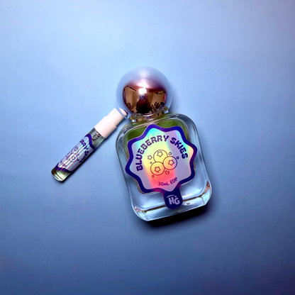 A clear glass perfume bottle with a metallic silver cap and a holographic blueberry skies label, shown next to a small matching sample vial on a blue gradient background.