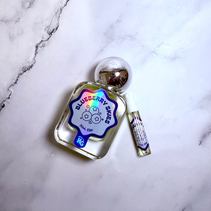 A clear glass perfume bottle labeled 'Blueberry Skies' with a holographic blue label and a small matching sample vial, placed on a white marble surface.