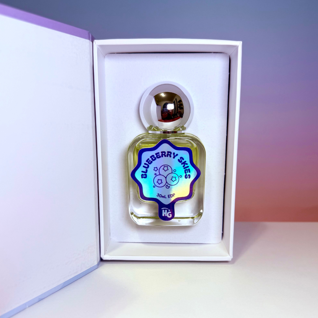 A small perfume bottle labeled 'Blueberry Skies' presented inside an open white gift box on a blue and peach gradient background
