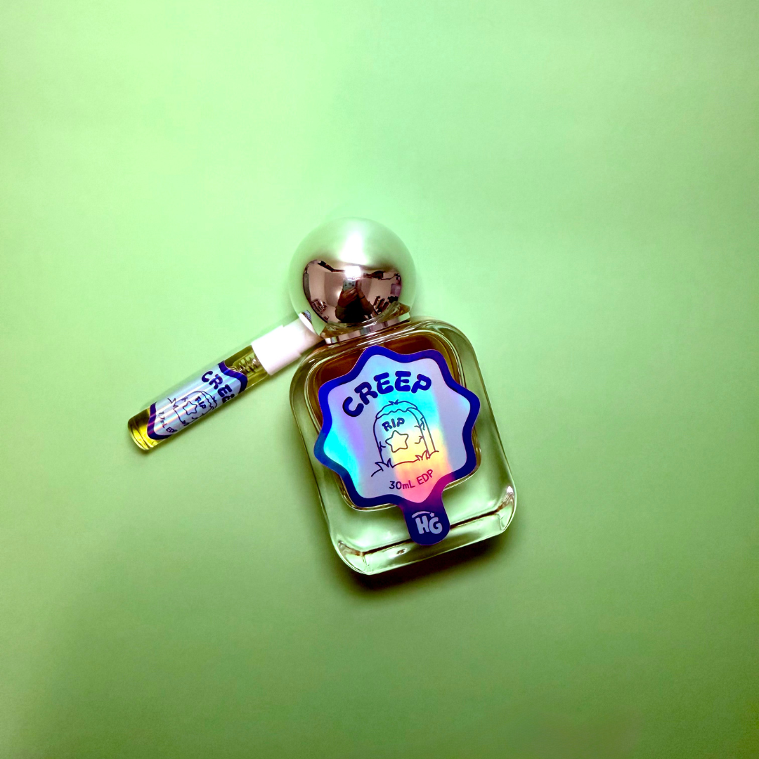 A small clear glass bottle with a reflective silver cap and a colorful holographic label reading 'CREEP' sits on a light green background, with a small sample vial resting beside it.