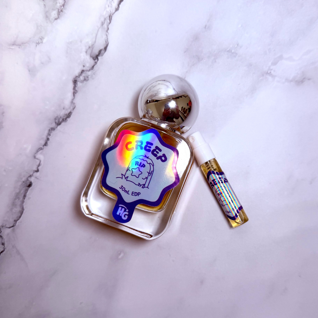Creep perfume bottle with a silver spherical cap and a small matching atomizer tube, placed on a white marble surface. The bottle label features a holographic rainbow sheen.