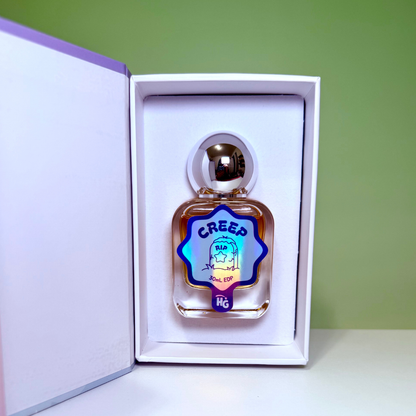 Creep perfume, displayed inside its white presentation box with a purple inner frame. The bottle is clear glass containing amber-colored liquid, featuring a metallic silver cap and a holographic blue label with the text 'CREEP' on a green background.