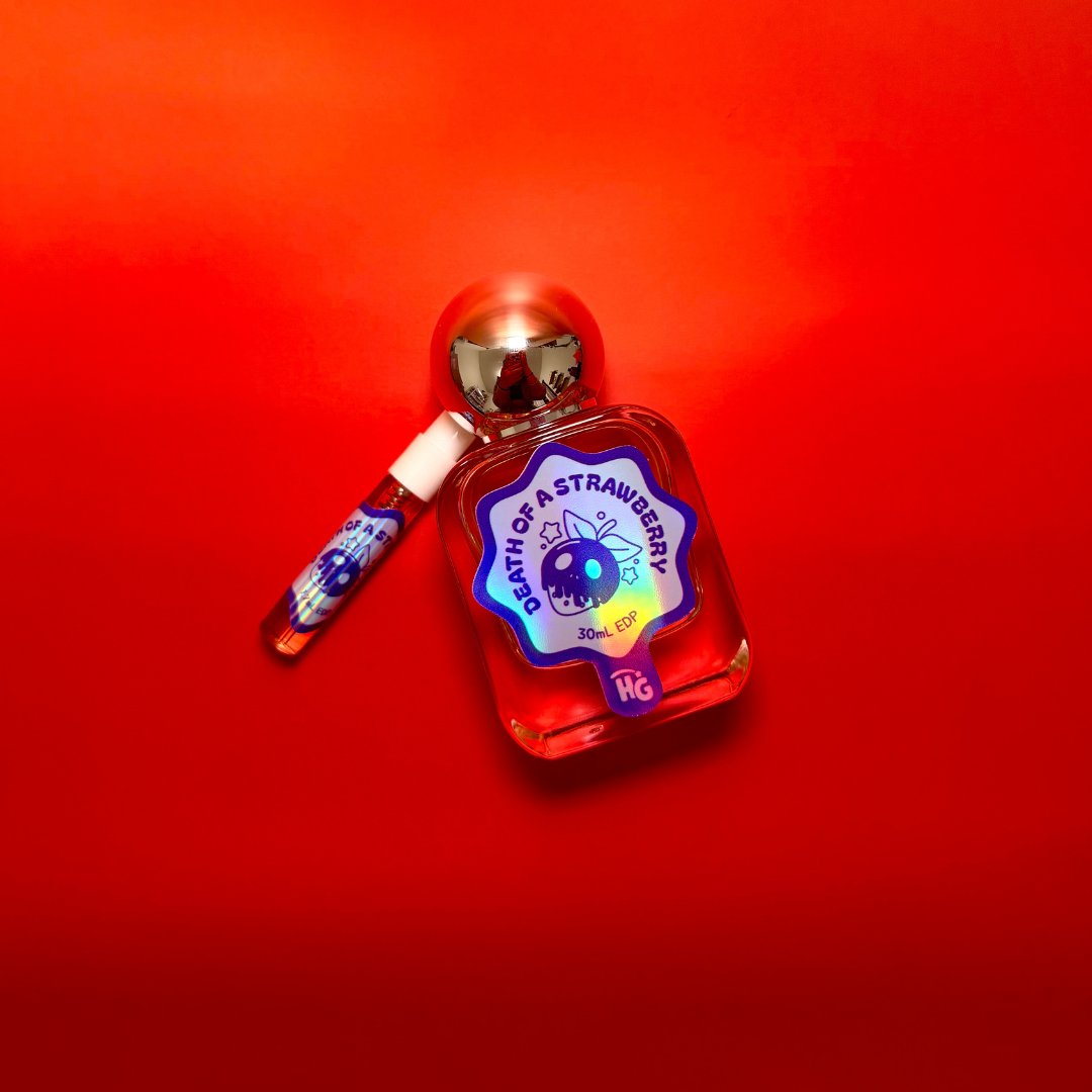 A small bottle of 'death of a Strawberry' perfume with a silver cap and a matching spray vial, set against a vibrant red gradient background.