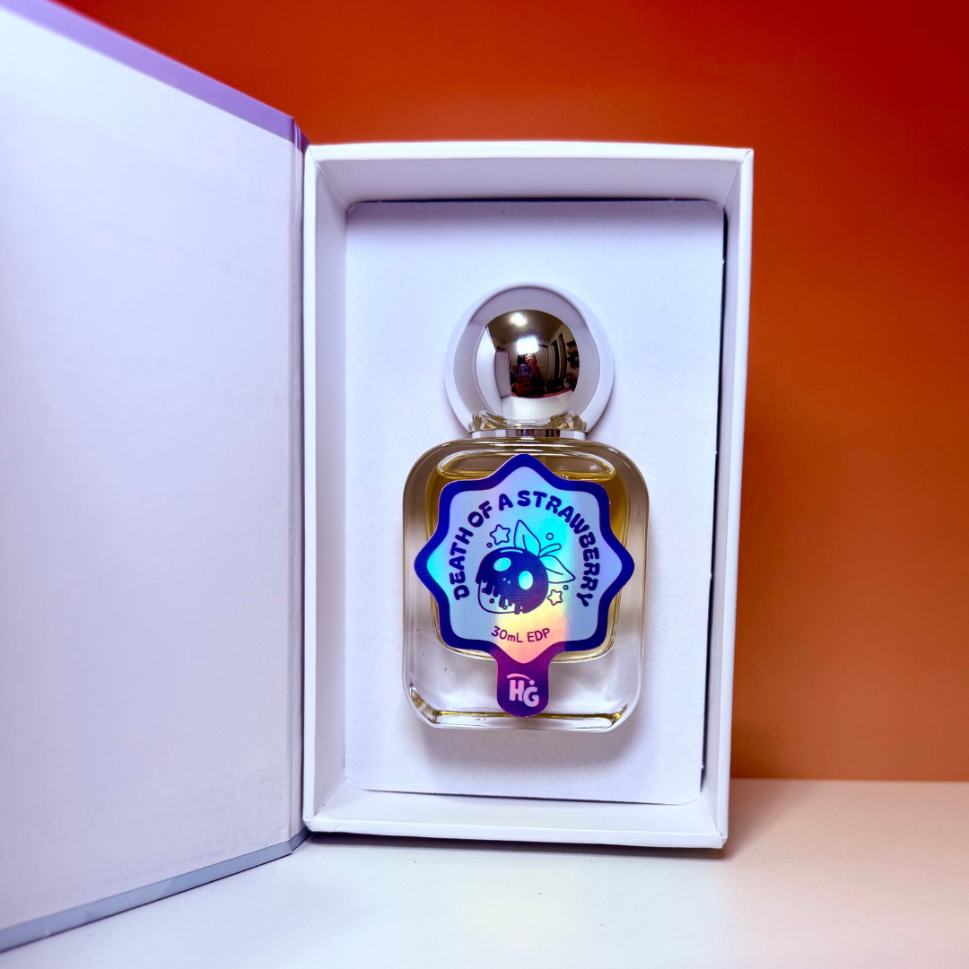 A small clear glass perfume bottle with a metallic silver cap, displayed inside an open white gift box. The bottle has a colorful holographic label reading 'DEATH OF A STRAWBERRY' on a red and orange gradient background.