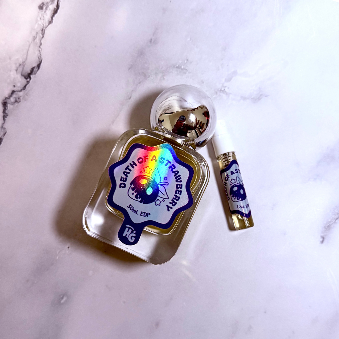 A small clear glass bottle with a clear cap and a blue, white, and holographic label reading 'DEATH OF A strawberry ' sits on a light marble surface, alongside a matching spray vial.