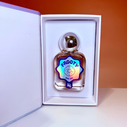 A clear glass perfume bottle with amber liquid, a silver cap, and a holographic blue label reading 'FROOTY' , displayed inside an open white gift box against an orange background.