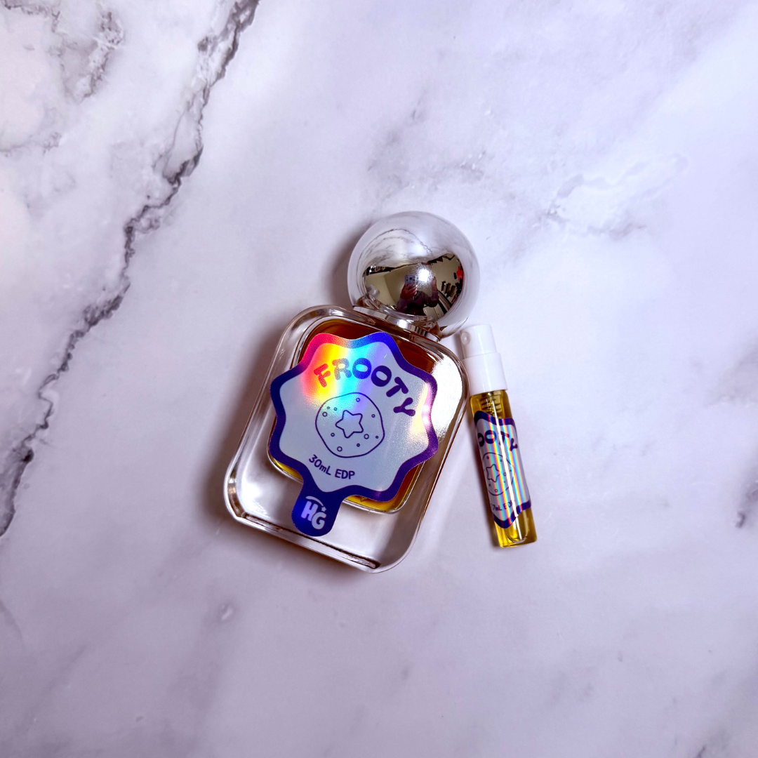 A small clear glass perfume bottle with a rounded silver cap and a matching travel spray vial, placed on a white marble surface. The bottle has a holographic flower-shaped label reading 'Frooty'.