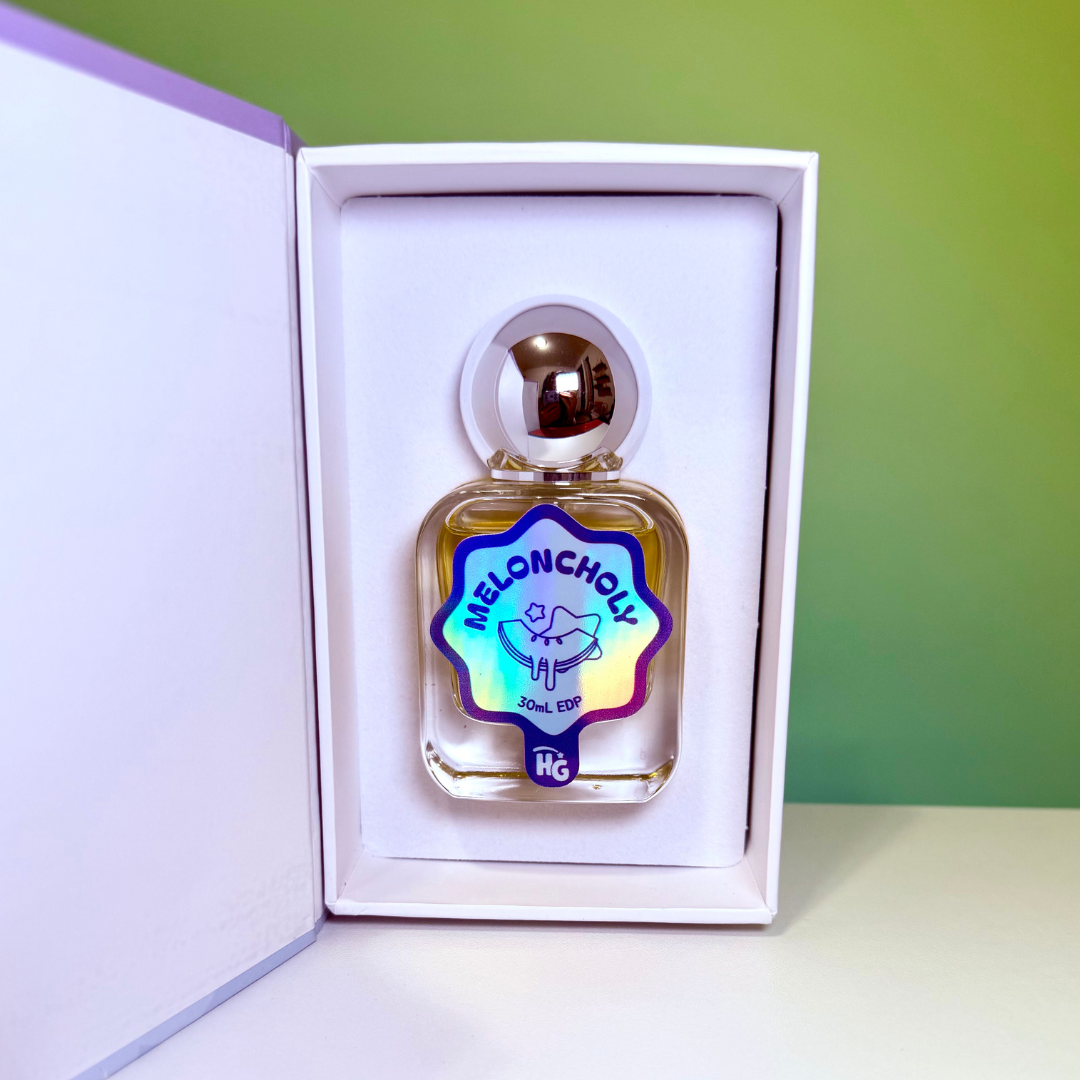 A perfume bottle labeled 'MELONCHOLY'  displayed inside an open white presentation box. The bottle has a clear glass body showing a golden-yellow liquid, a metallic silver cap, and a holographic blue label with a purple border.