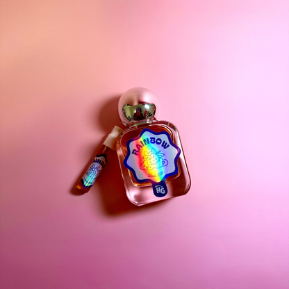 A small perfume bottle with a metallic pink cap and a colorful holographic label reading 'RAINBOW', shown next to a matching small vial on a pink gradient background.