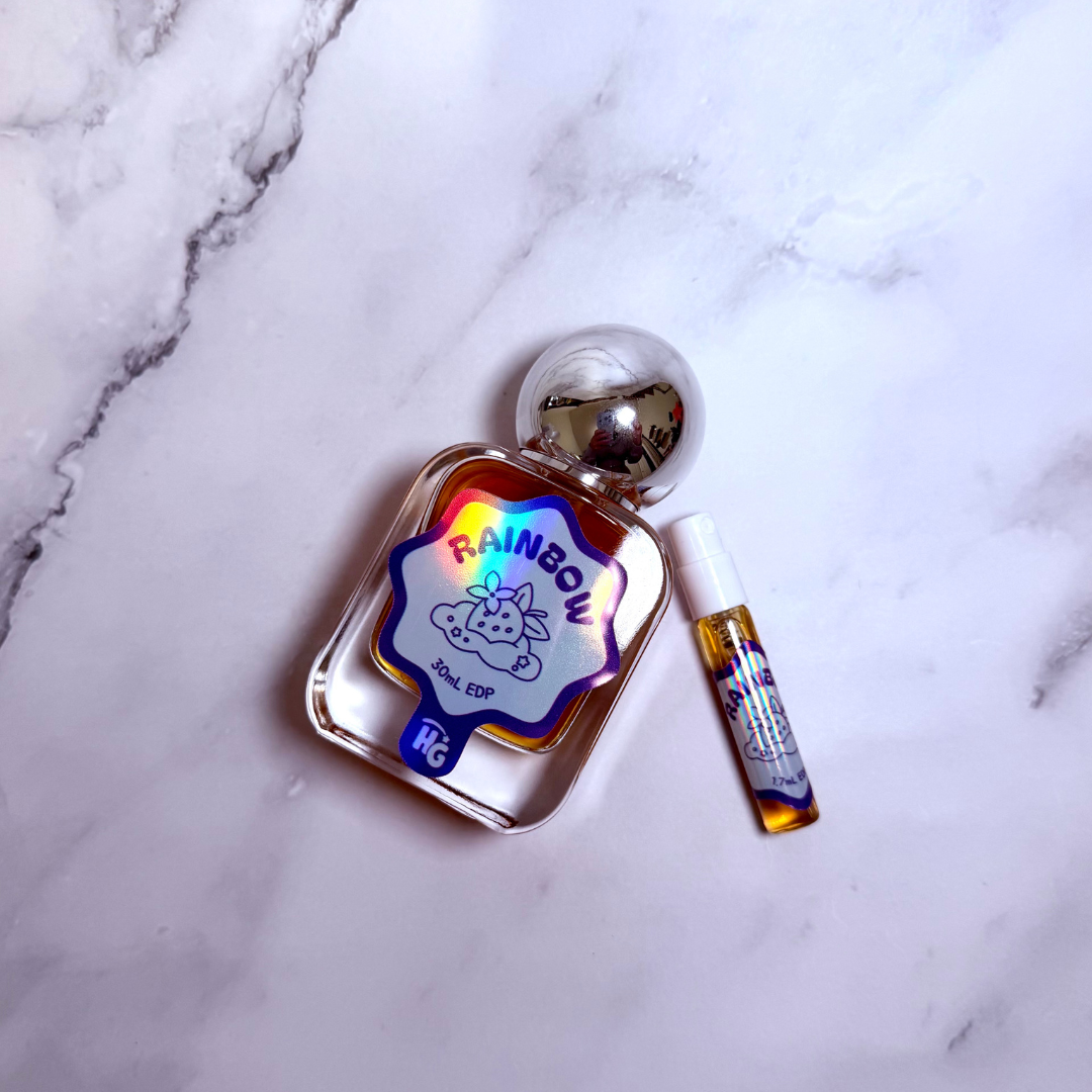 A clear glass perfume bottle labeled 'RAINBOW' with a colorful holographic label and a small matching sample vial, placed on a white marble surface.