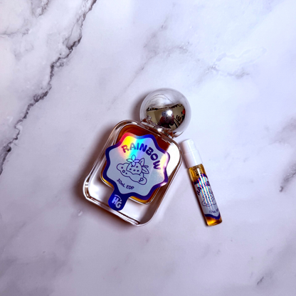 A clear glass perfume bottle labeled 'RAINBOW' with a colorful holographic label and a small matching sample vial, placed on a white marble surface.