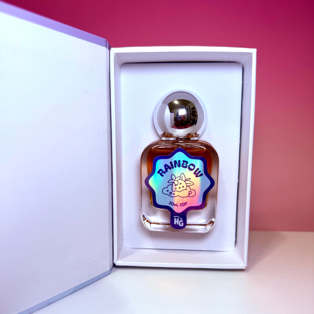 A small clear glass perfume bottle with amber liquid and a shiny silver cap, displayed inside an open white gift box. The bottle has a colorful holographic label reading 'RAINBOW', set against a pink gradient background.