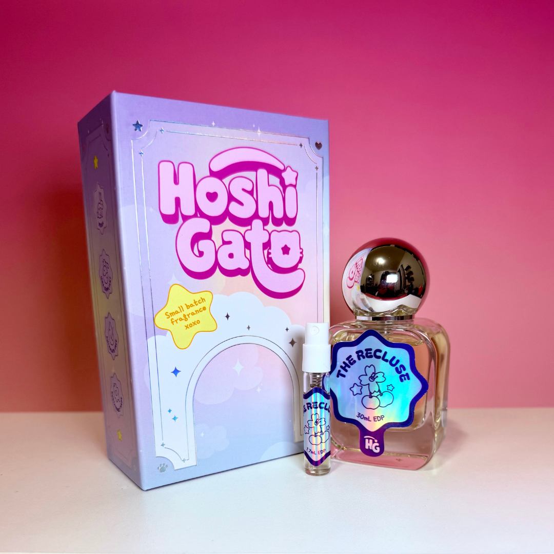 A pastel purple box, a small spray bottle, and a clear glass perfume bottle labelled "The Recluse" with a metallic cap, set against a pink gradient background.