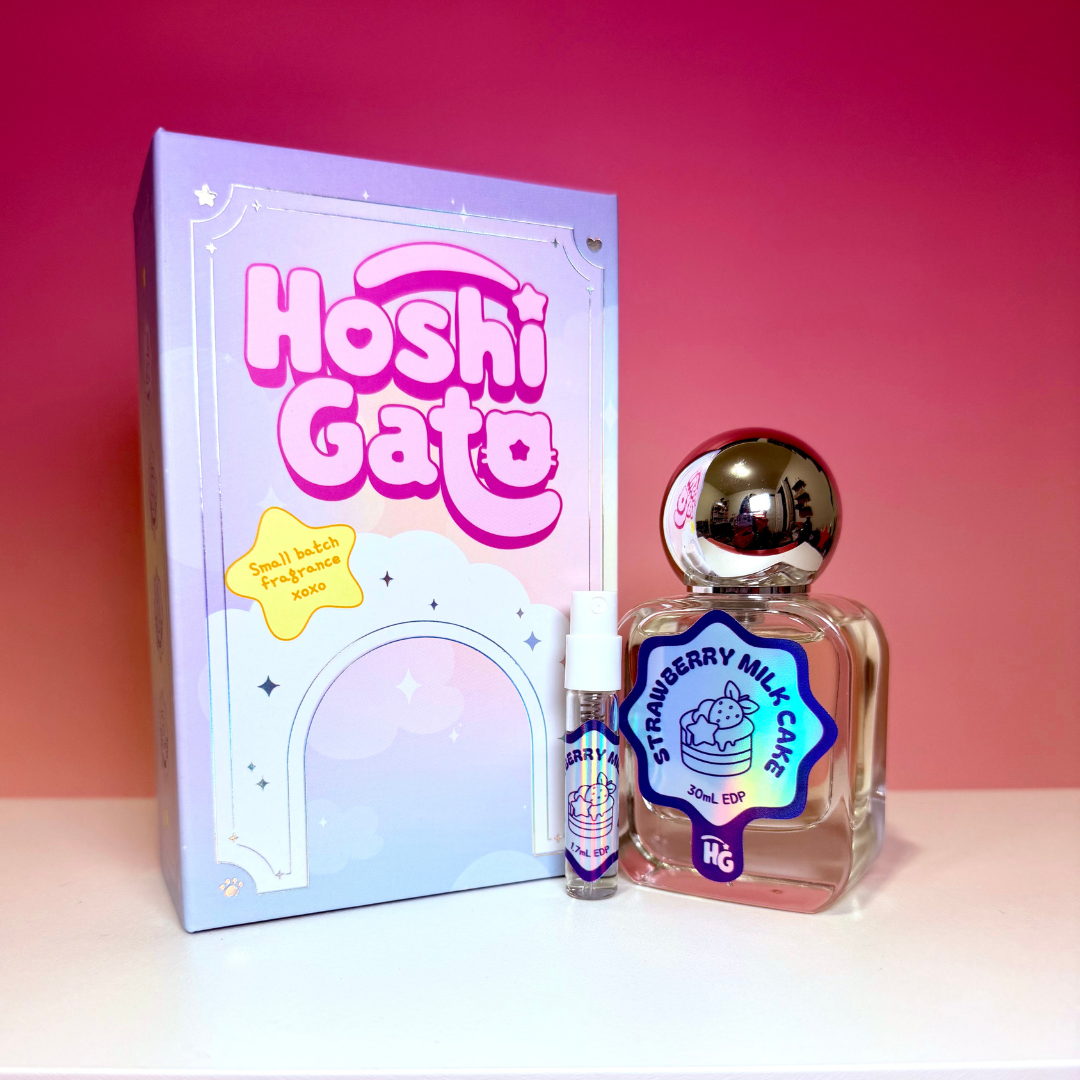 Hoshi Gato Strawberry Milk Cake including a lavender box, a clear glass bottle with a metallic silver cap, and a small travel spray vial, displayed on a white surface against a pink gradient background.