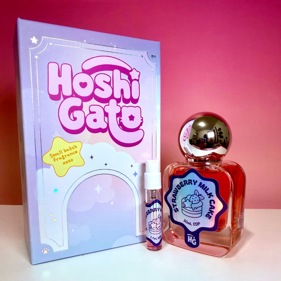 Hoshi Gato Strawberry Milk Cake including a lavender box, a clear bottle with red liquid, a metallic cap, and a small matching spray vial on a white surface against a pink gradient background.