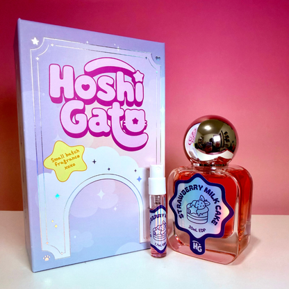 Hoshi Gato Strawberry Milk Cake including a lavender box, a clear bottle with red liquid, a metallic cap, and a small matching spray vial on a white surface against a pink gradient background.