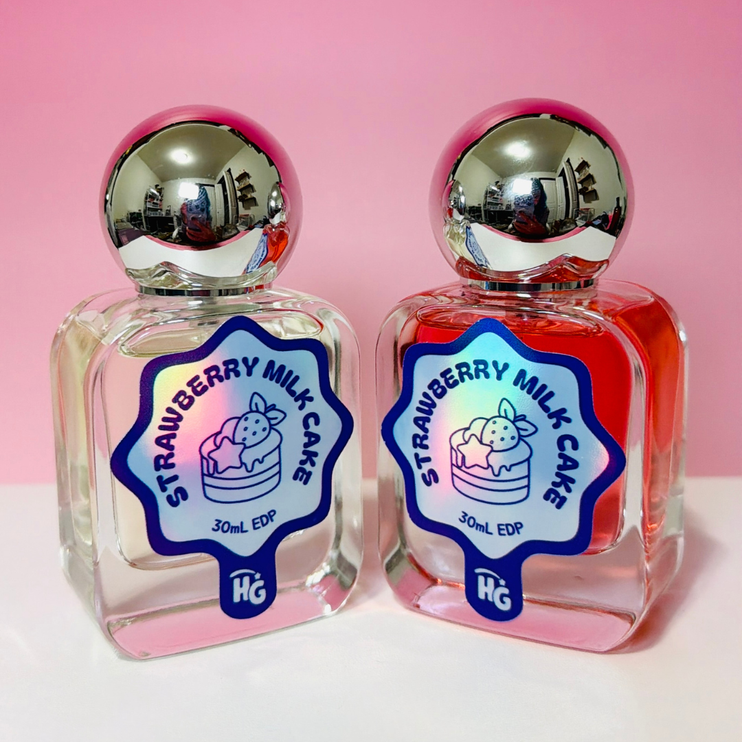 Two clear glass perfume bottles with shiny silver spherical caps, filled with clear and red liquids, labeled 'Strawberry Milk Cake' set against a pink background.