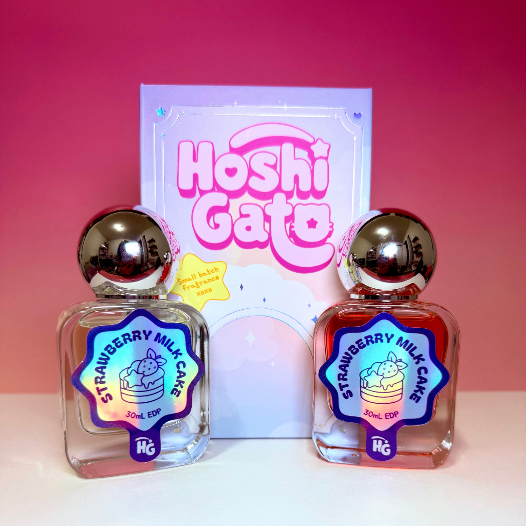 Two clear glass fragrance bottles labeled Strawberry Milk Cake, with metallic silver caps, displayed in front of a pastel pink and purple Hoshi Gato box on a pink gradient background.