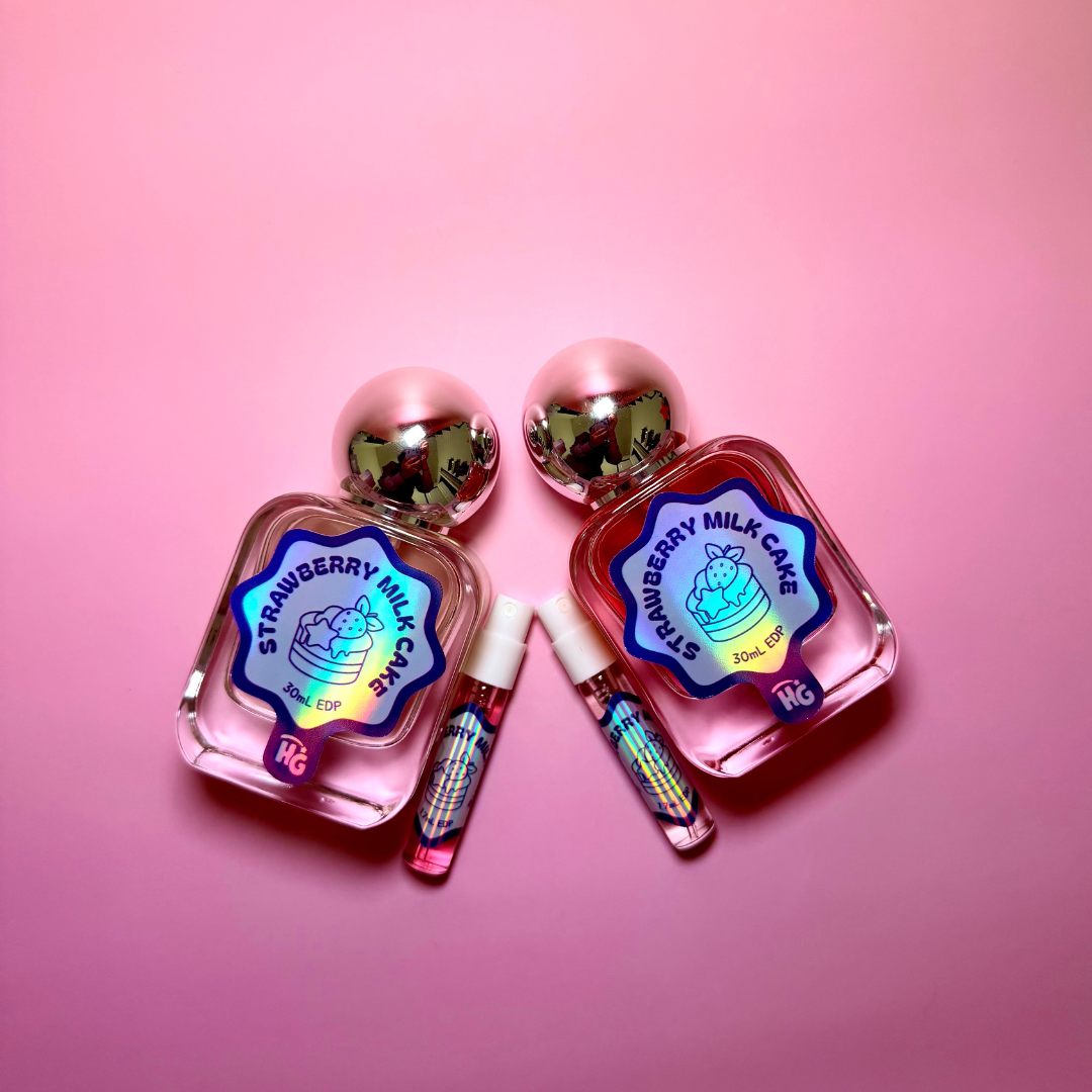 Two pink perfume bottles labeled 'Strawberry Milk Cake' with two matching sample vials, arranged on a solid pink background.