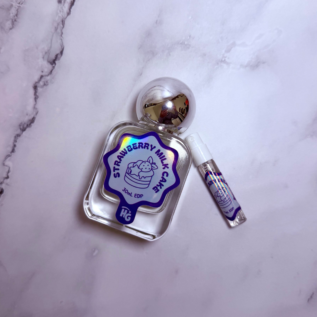 A clear glass perfume bottle labeled 'Strawberry Milk Cake' with a silver spherical cap, shown next to a small matching sample vial on a white marble surface.