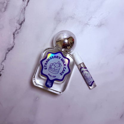 A clear glass perfume bottle labeled 'Strawberry Milk Cake' with a silver spherical cap, shown next to a small matching sample vial on a white marble surface.