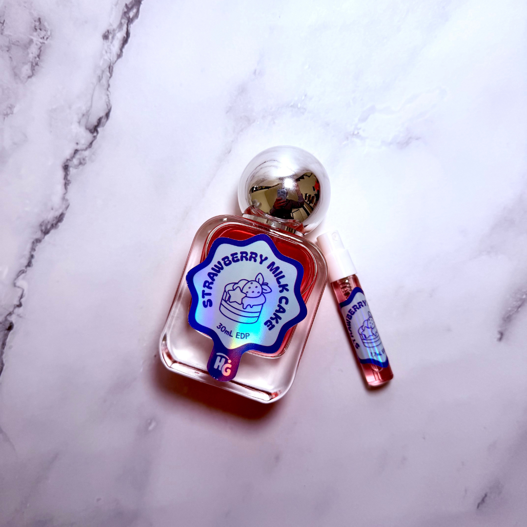 A clear glass perfume bottle labeled 'Strawberry Milk Cake' with red liquid, a silver spherical cap, shown next to a small matching sample vial on a white marble surface.