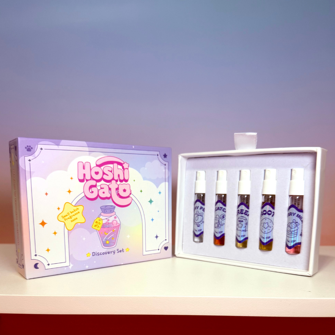 A Hoshi Gato discovery set shown with its illustrated box on the left and the open tray containing five small spray vials on the right, placed on a shelf against a soft blue-to-pink gradient background.