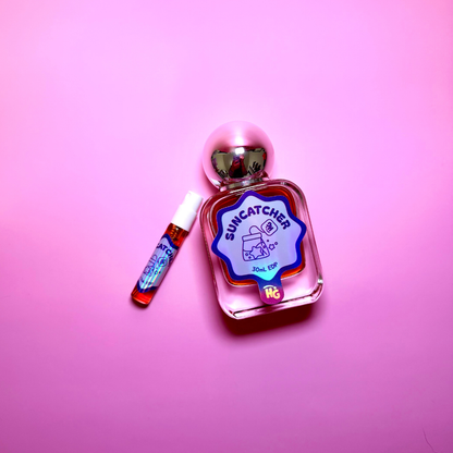 A clear glass perfume bottle labeled 'Suncatcher' with a pink and blue label, shown next to a small matching spray vial on a solid pink background.