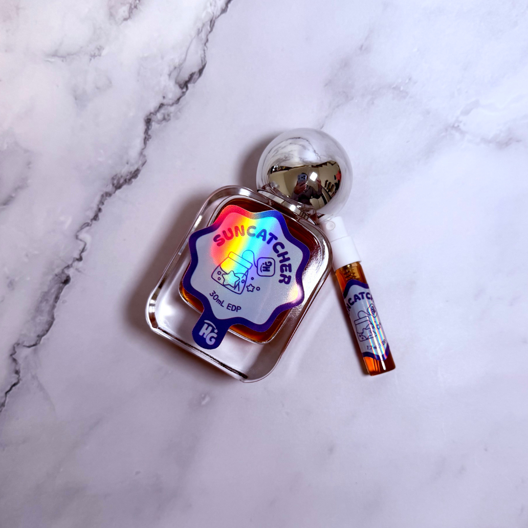 A small clear bottle labeled 'Suncatcher' with a holographic rainbow sticker and a matching small tube placed beside it on a white marble surface.