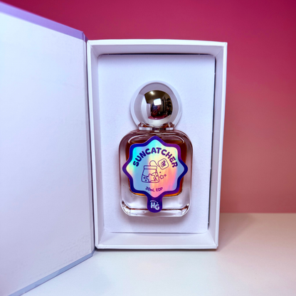 A small clear glass perfume bottle labeled 'SUNCATCHER' with a holographic blue-purple label, displayed inside an open white gift box against a pink gradient background.
