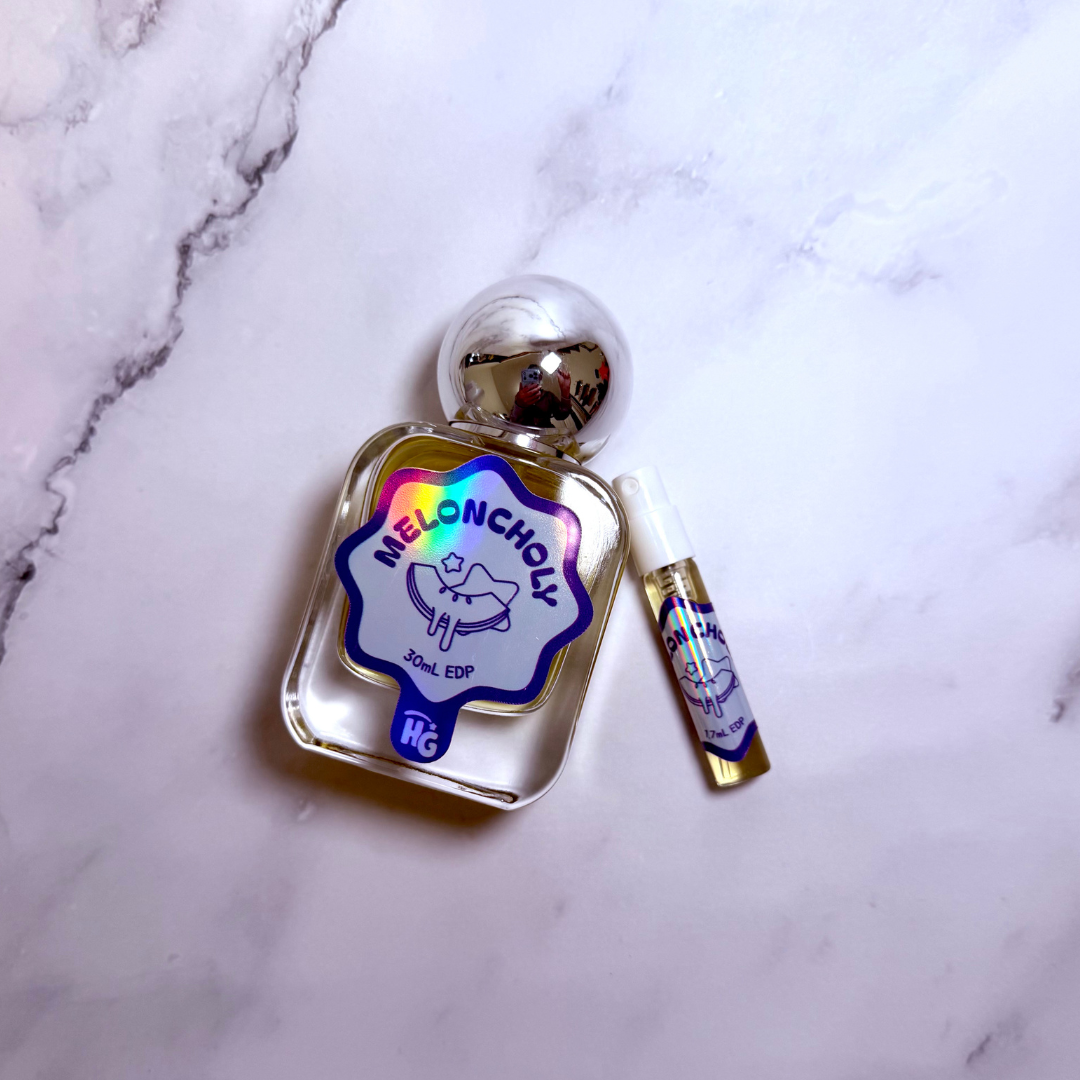 A small perfume bottle labeled 'Meloncholy' with a silver cap and a matching travel spray vial, placed on a white marble surface.