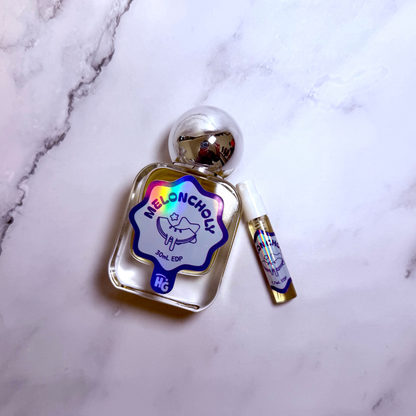 A small perfume bottle labeled 'Meloncholy' with a silver cap and a matching travel spray vial, placed on a white marble surface.
